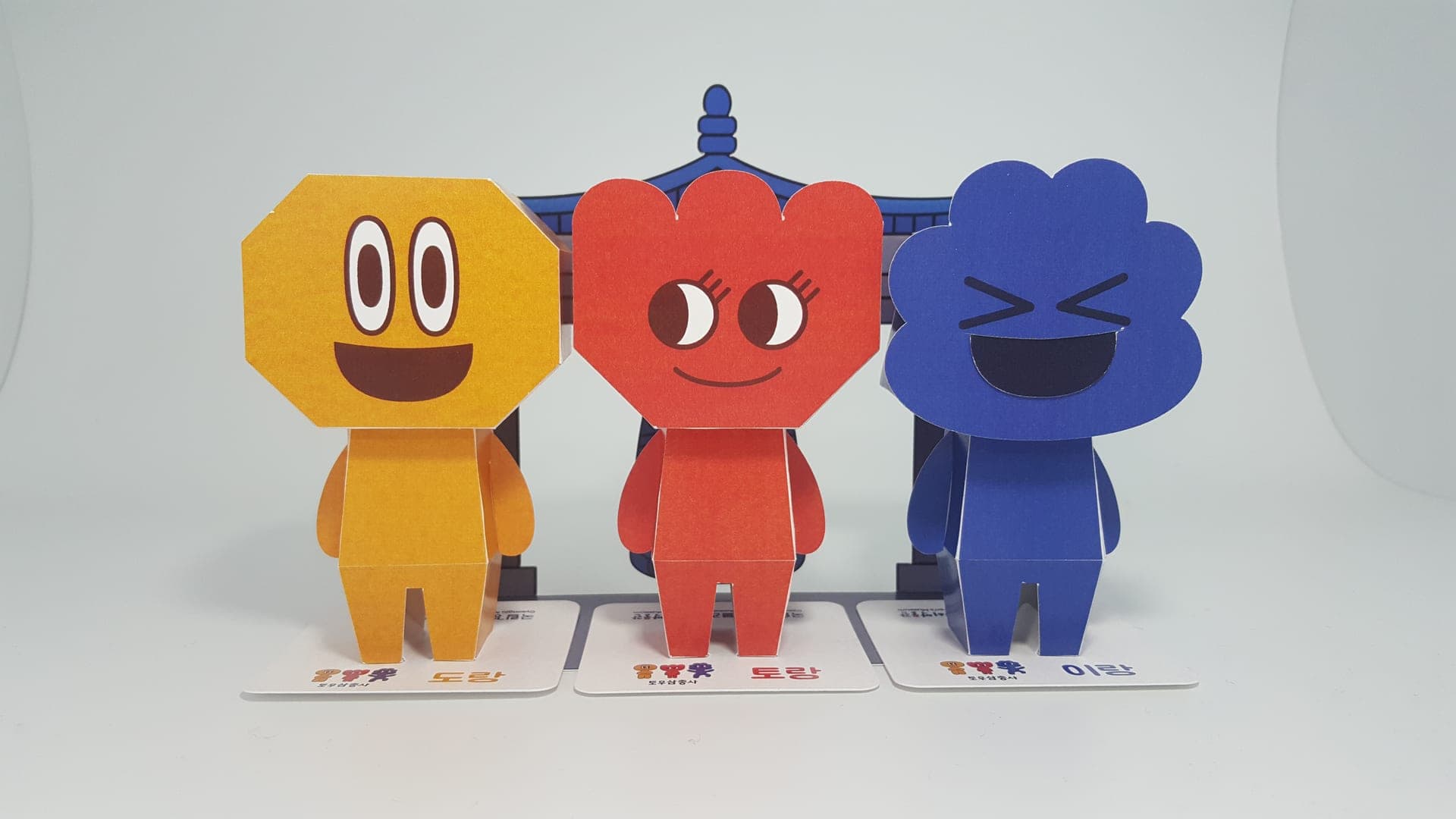 노란 도랑, 빨간 토랑, 파란 이랑이 이름표 받침 위에 나란히 서고 뒤로 파란 기와 종각이 펼쳐진 도토리 페이퍼토이